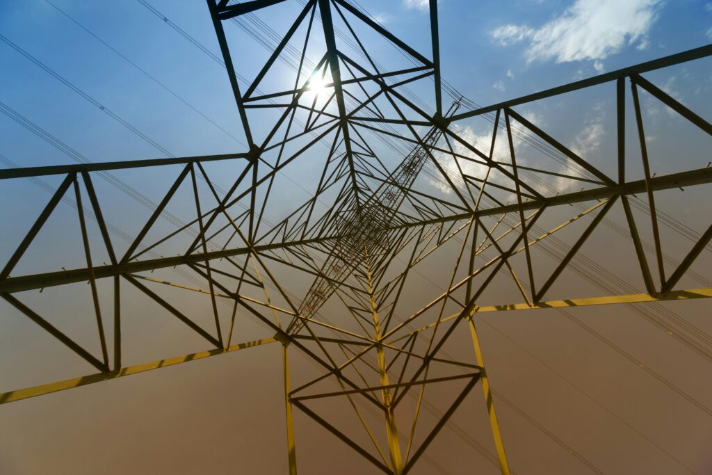 Looking up view of a steel transmission tower with sun glare and blue sky.